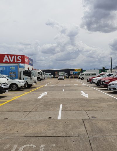 Directional arrows on the roadway of an Avis Car Rental site to direct traffic around the site. Photo also includes car parks in white on the right side and yellow on the left side, and a pedestrian crossing.