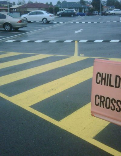 A carpark showing a yellow pedestrian crossing with a Children Crossing sign and directional arrows around the carpark.