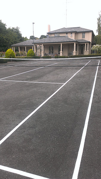 White tennis court markings on a black surface in the yard of a house.